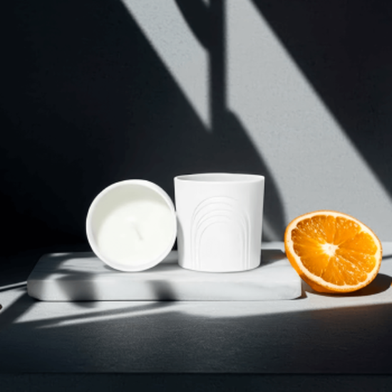 Composition on white marble: two white candle jars (one standing arch pattern, one lying white wax). Right, a juicy half-orange. Dramatic geometric shadows cross scene, creating striking contrast.