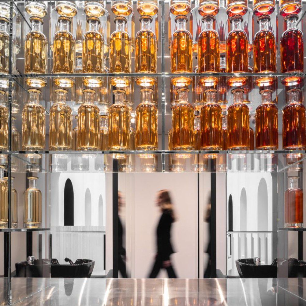 A modern salon wall display features glass shelves with colorful bottles (gold to red) reflecting light. Mirrored surfaces below show two black washing sinks and a blurred silhouette. The overall ambiance is sophisticated.