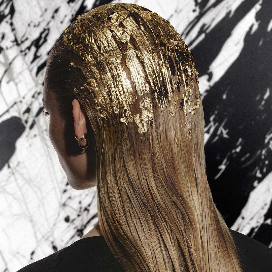 A blonde woman seen from behind, wearing gold leaves in her hair and a black Carita dressing gown.