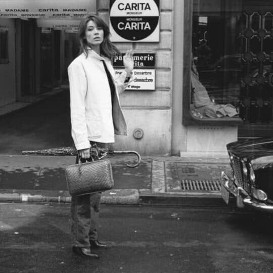A woman in a light coat stands in a narrow street, looking back towards a sign that reads "CARITA." The image is in black and white, capturing a vintage aesthetic.