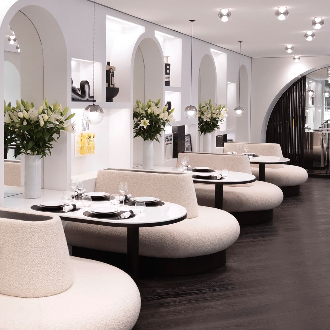 Elegant dining area with curved banquettes, set round tables, arched white walls with alcoves, and white lily bouquets. Chic, modern ambiance.