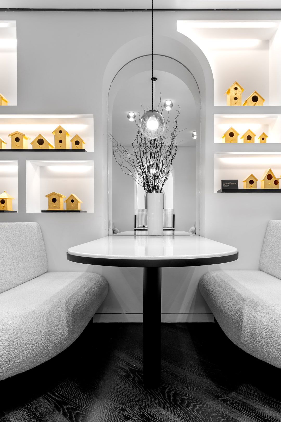 A modern interior featuring a round table with a black leg, surrounded by two curved white sofas. Behind the table, there is a mirror and shelves displaying yellow birdhouses against a white backdrop, complemented by a decorative branch in a vase. The space is well-lit and minimalistic.