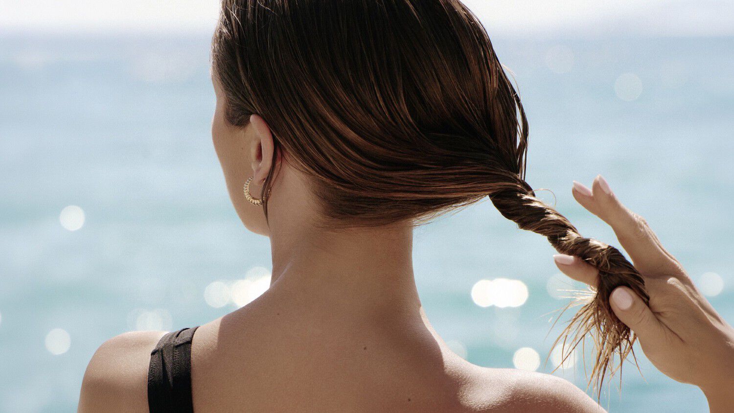 A woman seen from behind at the water’s edge, running her hands through her hair.
