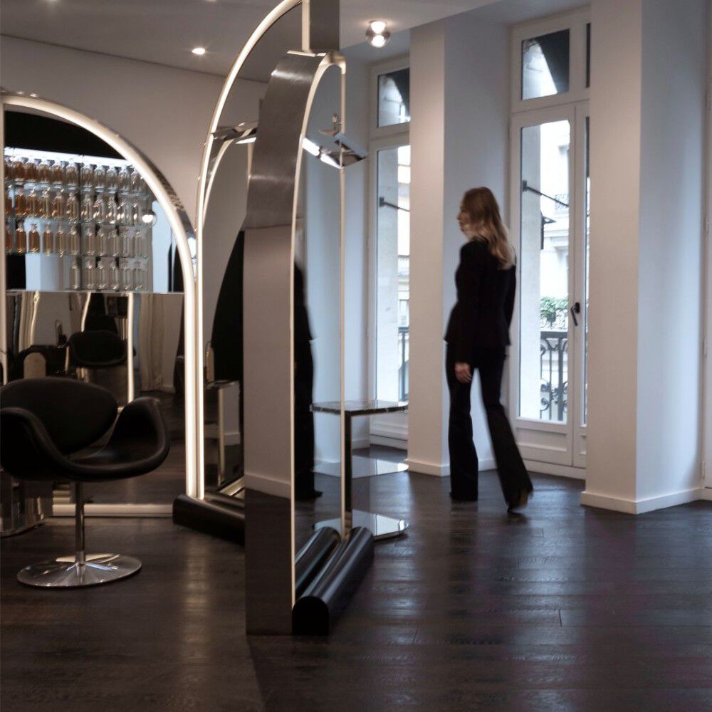 An elegant, modern hair salon. Large, arched, reflective structures feature integrated lighting. A sleek black chair is visible on the left. Wall shelves display bottles. A woman walks towards windows with a Parisian balcony.
