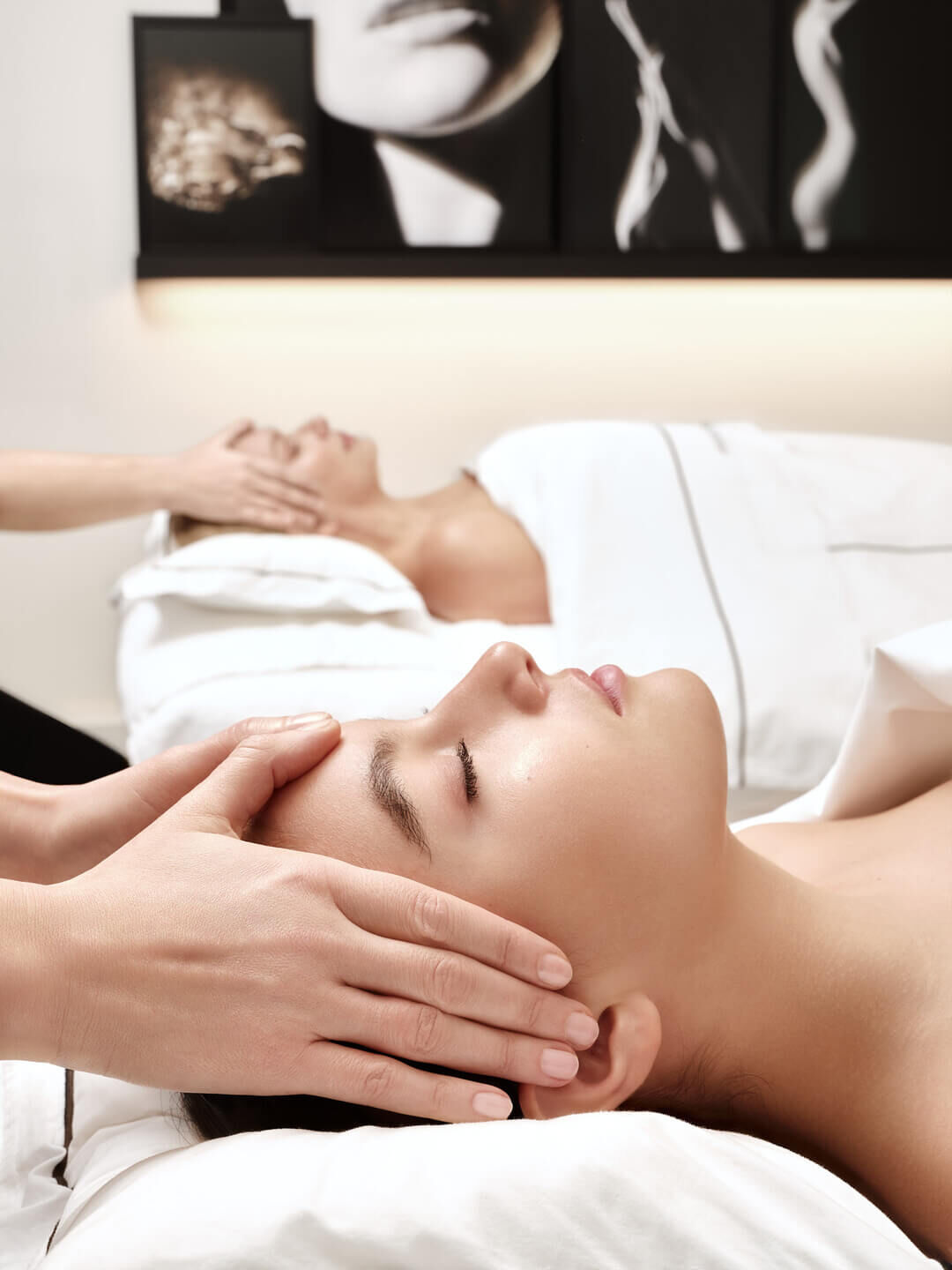 Two women relax, eyes closed, receiving soothing head and facial massages in a serene spa setting, emphasizing tranquility and well-being.