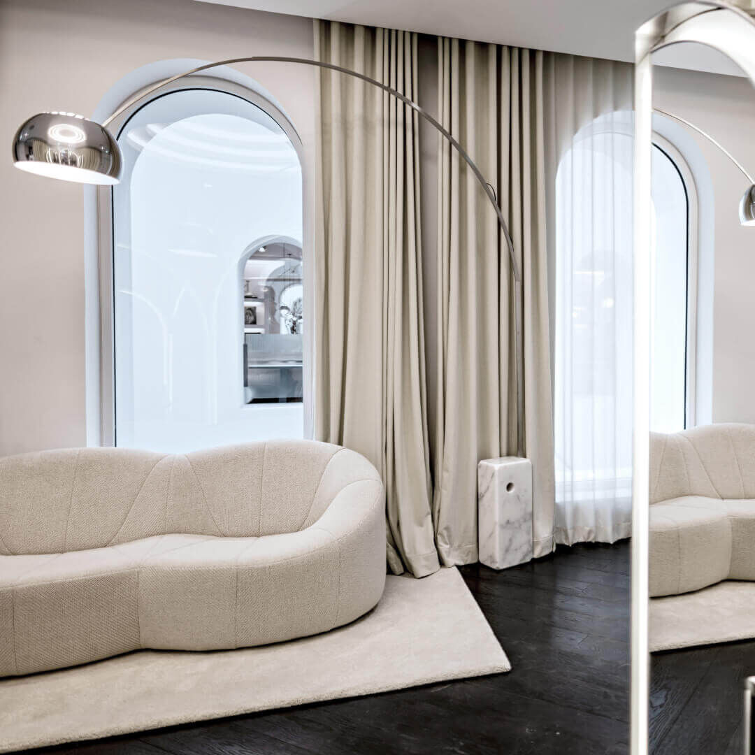 Modern lounge area. A beige, organic-shaped sofa rests on a matching rug, in front of arched windows with beige curtains. An arc lamp illuminates it. A vertical mirror reflects the scene, adding spaciousness and brightness.