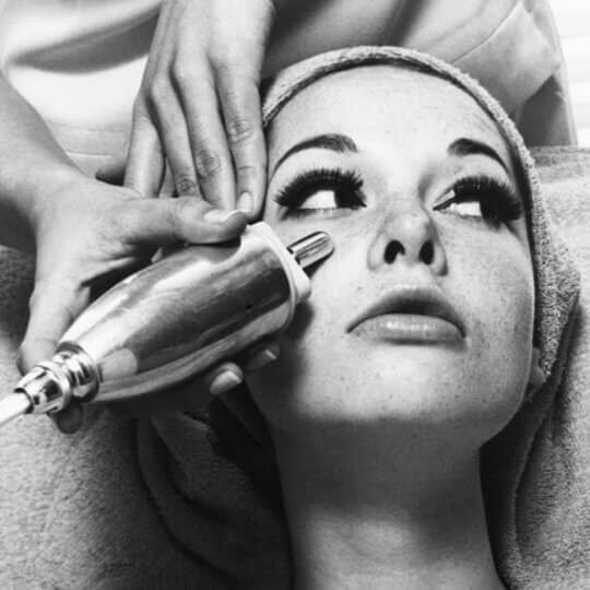 A close-up of a woman undergoing a skin treatment, with a handheld device positioned near her face while a therapist's hand adjusts the device. The woman has long eyelashes and is wearing a towel on her head.