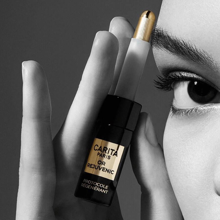 A close-up of a person holding a sleek black and gold cosmetic product near their face, with a focus on their expressive eyes against a dark background.