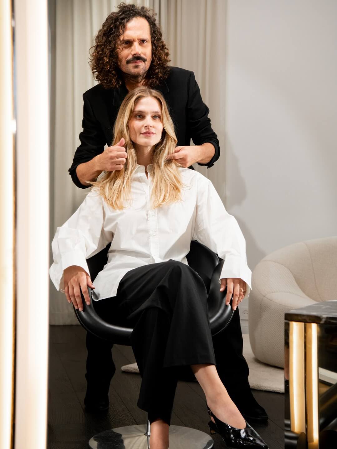 A master stylist, with a confident gesture, perfects a client's luminous blonde hair, embodying bespoke luxury and exceptional salon craftsmanship.