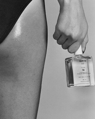Black and white close-up: A hand holds a Carita Paris "Fluide 14" bottle next to a person's moisturized upper thigh, highlighting skin nourishment.