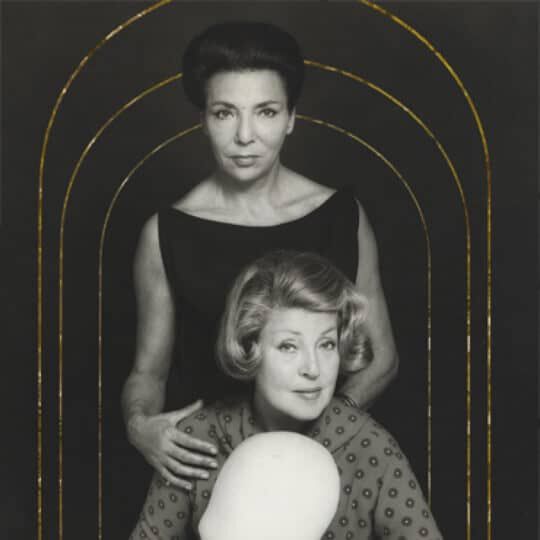 A black and white image of two women in elegant attire, one standing behind the other, framed by an arch with golden lines. The background is dark, emphasizing the figures in the foreground.