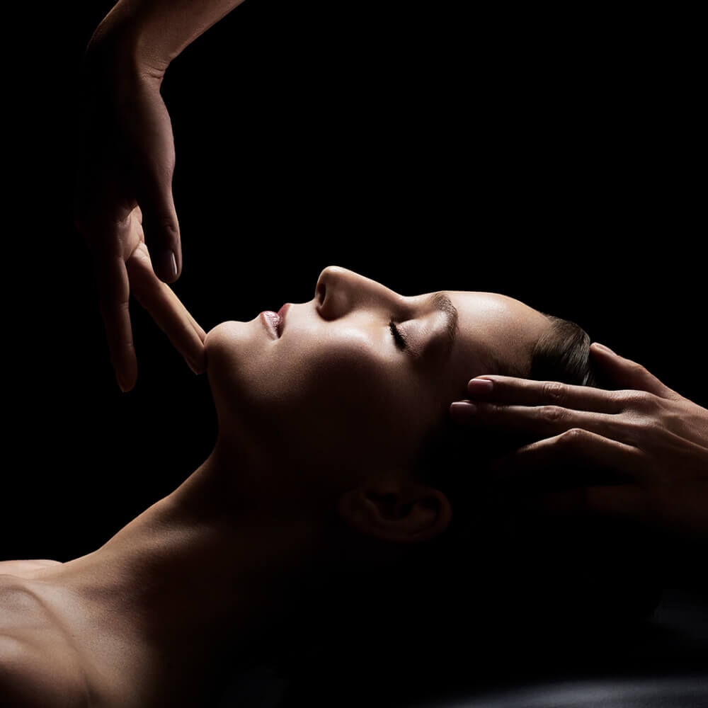 A close-up image of a woman with her eyes closed, receiving a facial massage, with hands gently positioned on her face, set against a dark background.