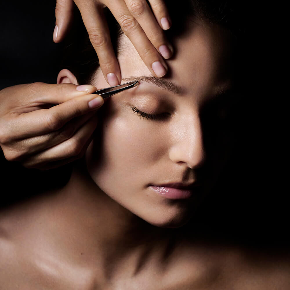 A close-up of a person's face with one eye closed, as a hand uses tweezers to shape their eyebrow against a dark background.