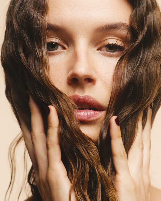 Close-up of a woman with wavy brown hair, framing her face with her hands against a light beige background.