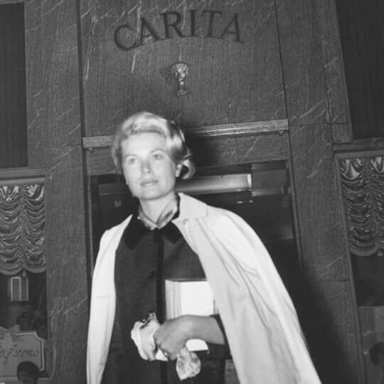 A black and white portrait of a woman with light-colored hair, standing in a stylized indoor setting.
