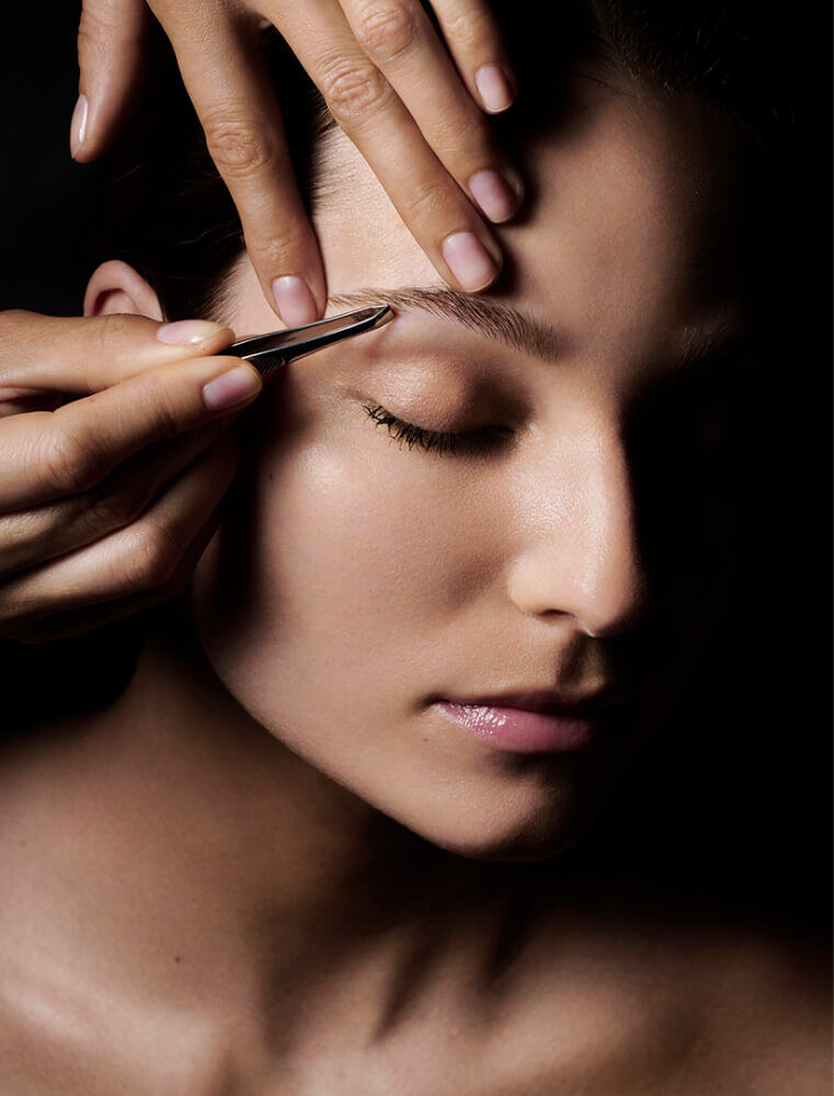 Close-up of hands using tweezers to shape a woman's eyebrow.