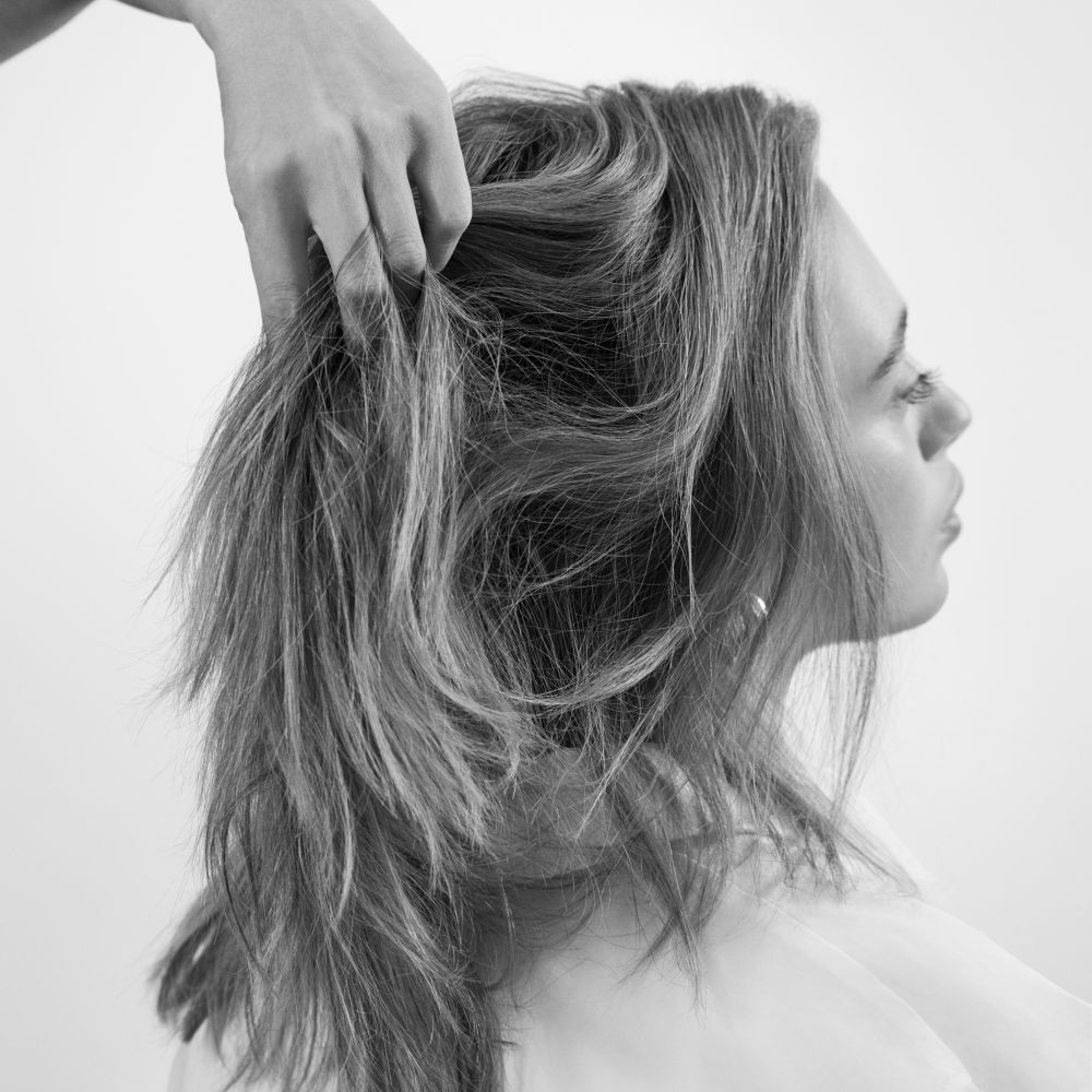 Monochrome close-up of a woman in profile, her hair being styled or massaged by a hand, highlighting volume and texture.