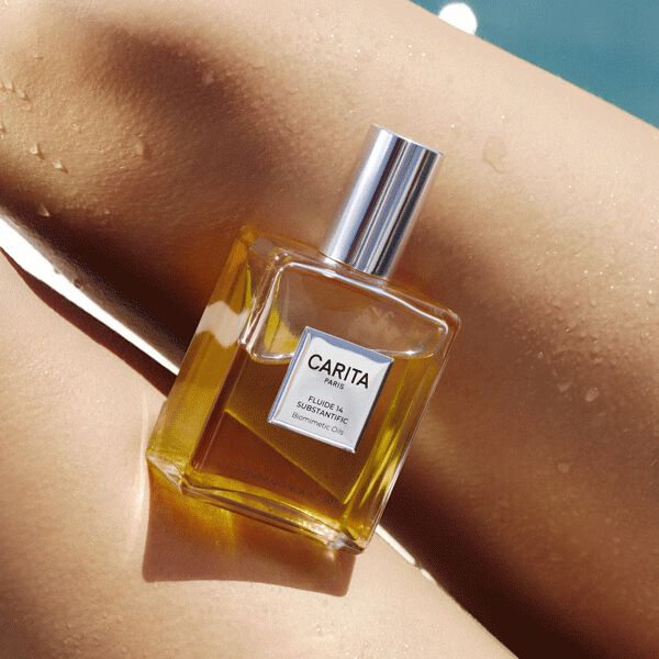 A glass bottle of Carita Fluide 14 dry oil resting on sun-kissed, wet skin with water droplets, with a blue swimming pool reflecting sunlight in the background.​