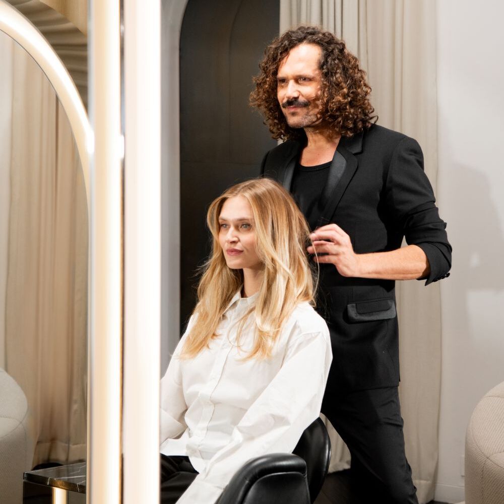 In a modern salon, a blonde woman in a white shirt sits in a black chair, looking left. A man in a black suit adjusts her hair. The scene is framed by mirrors, reflecting light beige curtains and a dark arched doorway.