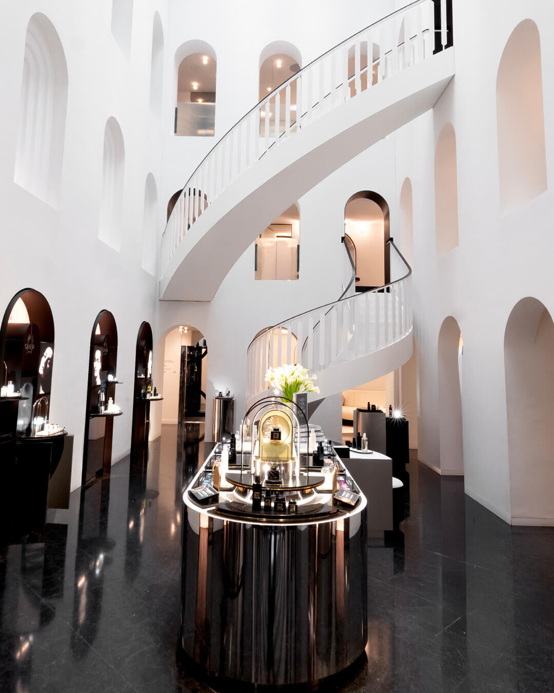 Modern, luxurious boutique interior featuring a large white spiral staircase, arches, and product displays on glossy black floors.