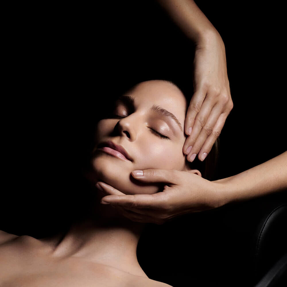 A close-up of a woman with her eyes closed, enjoying a facial massage as one hand gently holds her jaw while the other supports her head, against a dark background.