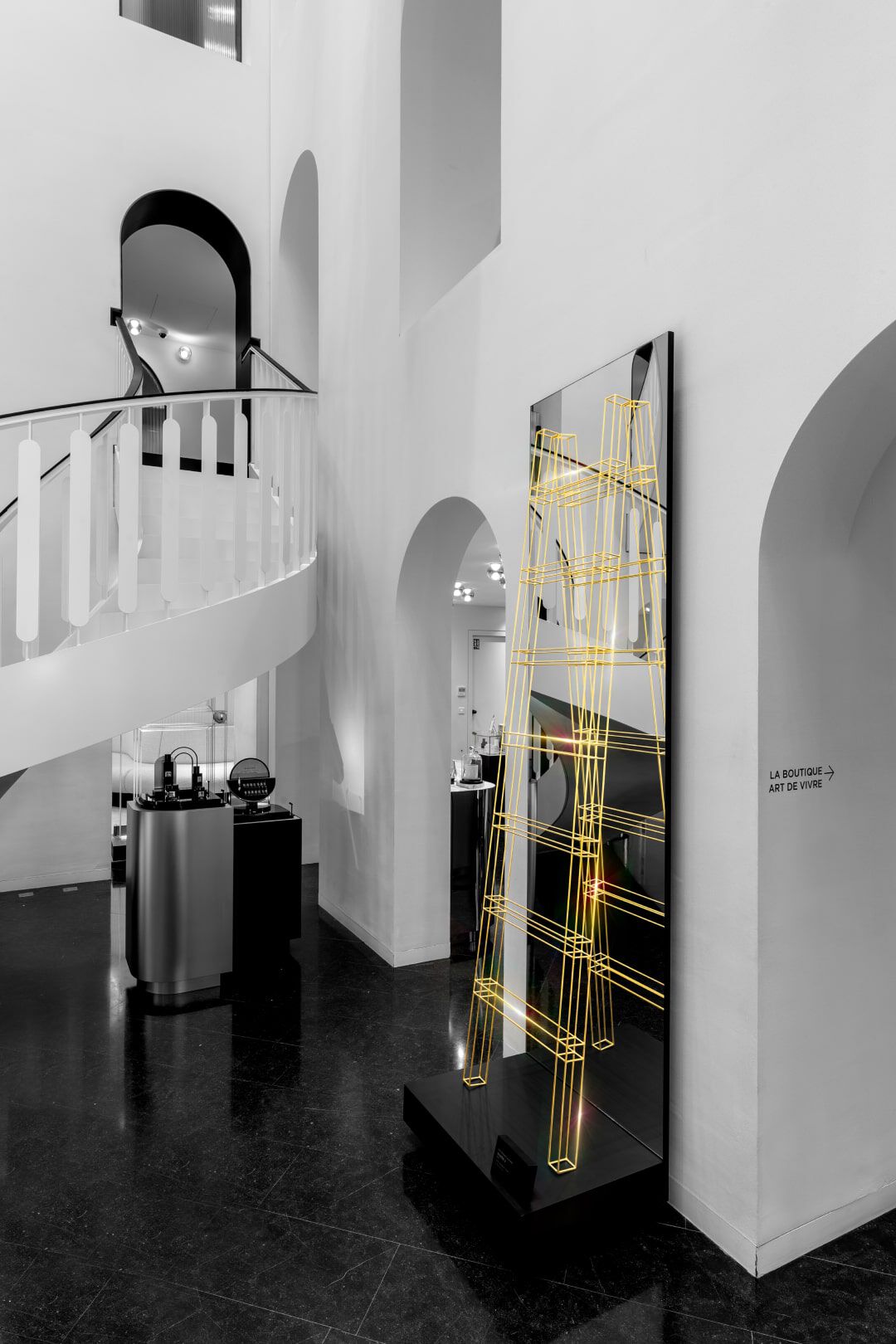 A modern interior space featuring a staircase and a dark floor, adorned with a sleek mirror displaying a yellow geometric design. The surroundings are minimalist with white walls and artistic elements.