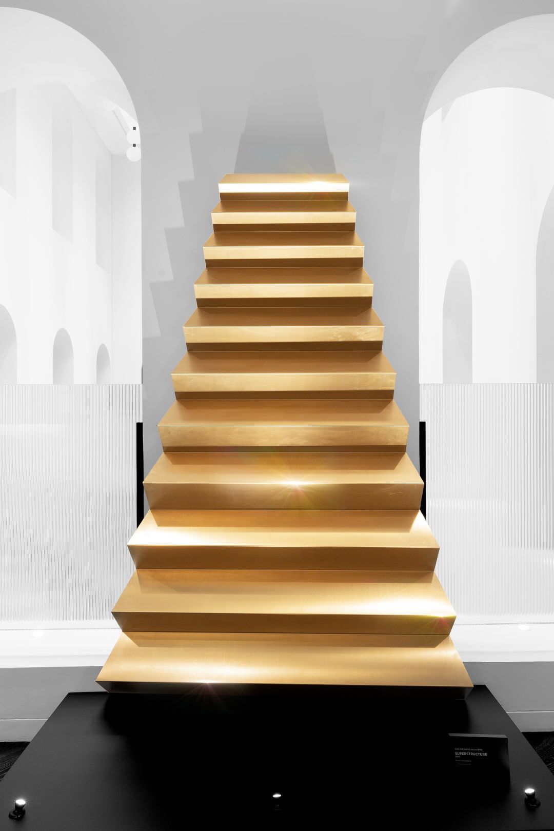 A golden staircase made of layered steps, displayed against a minimalist white background in a modern gallery setting.