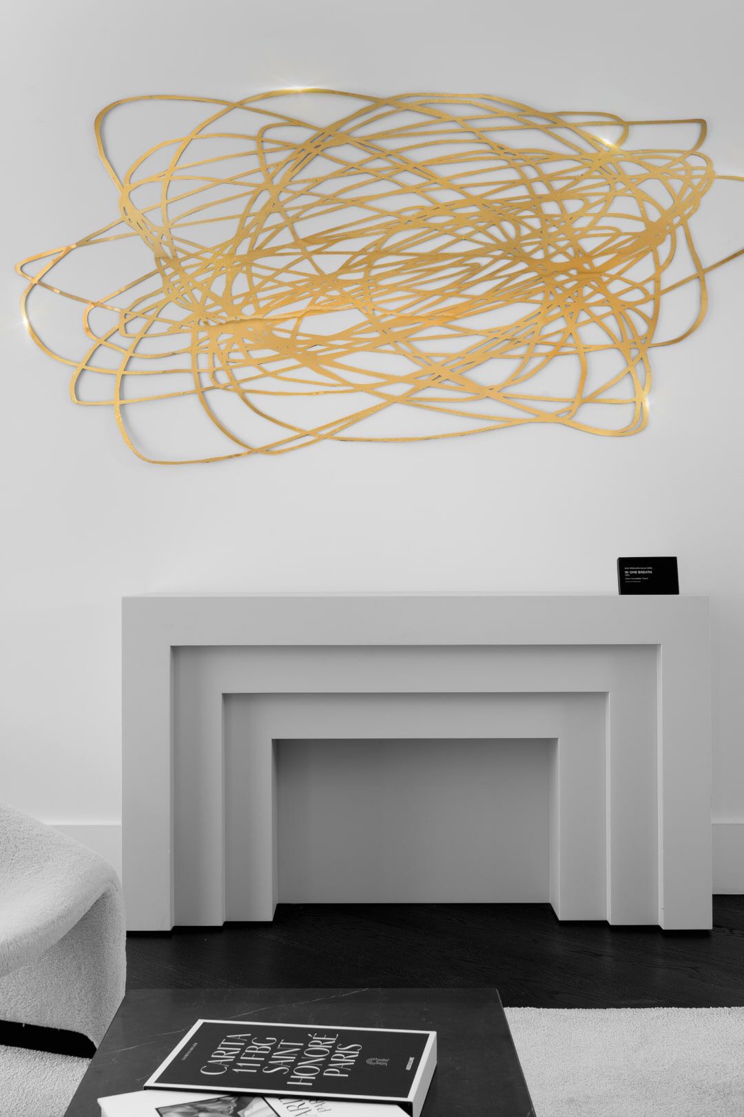 A modern, abstract gold wall sculpture is displayed above a white mantelpiece, which is styled with a black book and an informational card on a small stand. The background is a simple white wall.
