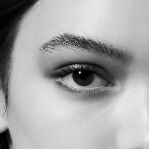 Intense monochrome close-up of a female eye, highlighting voluminous lashes and a defined eyebrow, with a bright catchlight in the iris.