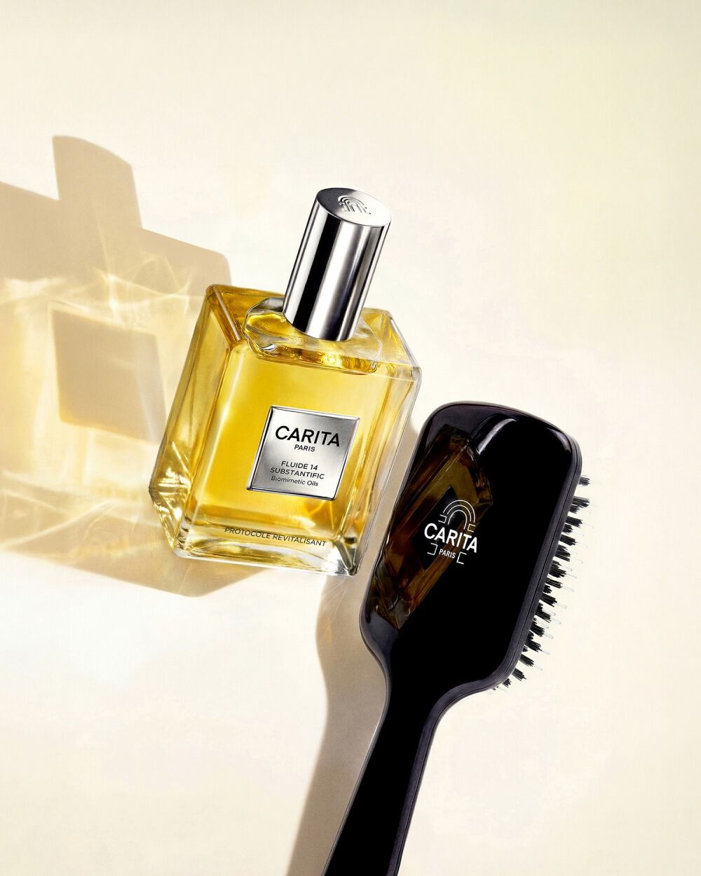 A bottle of golden Carita Fluide 14 and a sleek black Carita hairbrush on a light neutral background with soft light reflections.