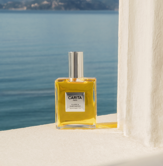 A square bottle of Fluide 14 dry oil, with its cap and its plaque on top. It is placed on a window sill without glass, with mountains and the sea in the background, evoking summer.​