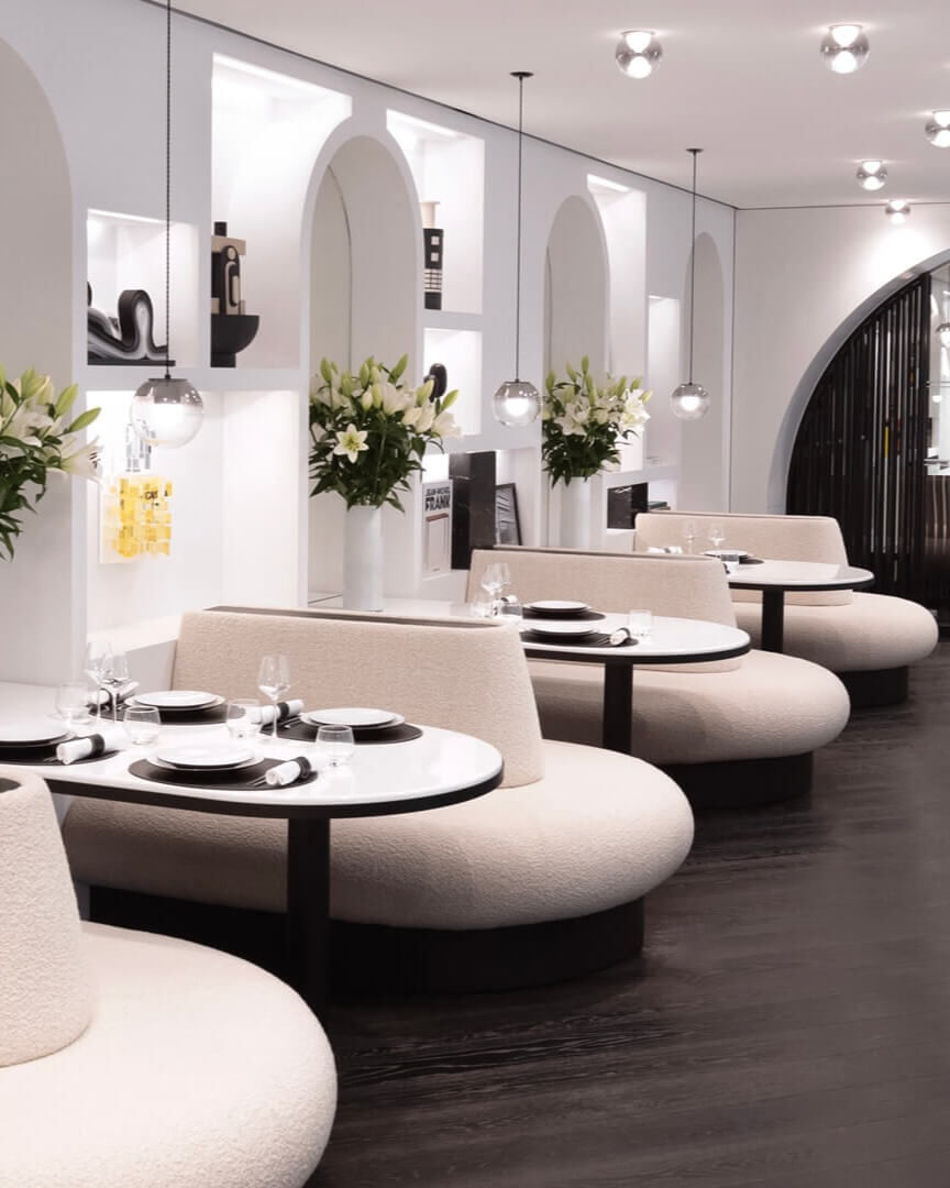 Elegant dining area with curved banquettes, set round tables, arched white walls with alcoves, and white lily bouquets. Chic, modern ambiance.