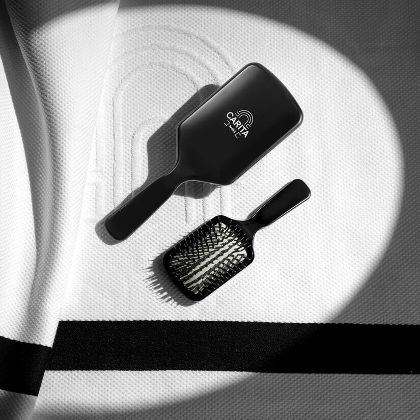 Two black hairbrushes, one large and one smaller, are laid on a textured white surface, possibly a terry cloth or uniform. The larger brush, located at the top left, displays the CARITA Paris logo with the arch motif. The smaller brush is positioned at the bottom right. Crisp shadows are visible on the surface.