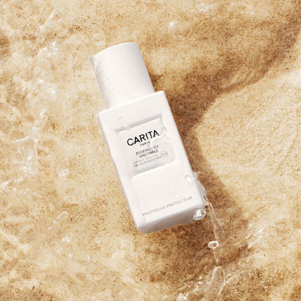 A white bottle of Carita’s Essence UV sitting in the sand.