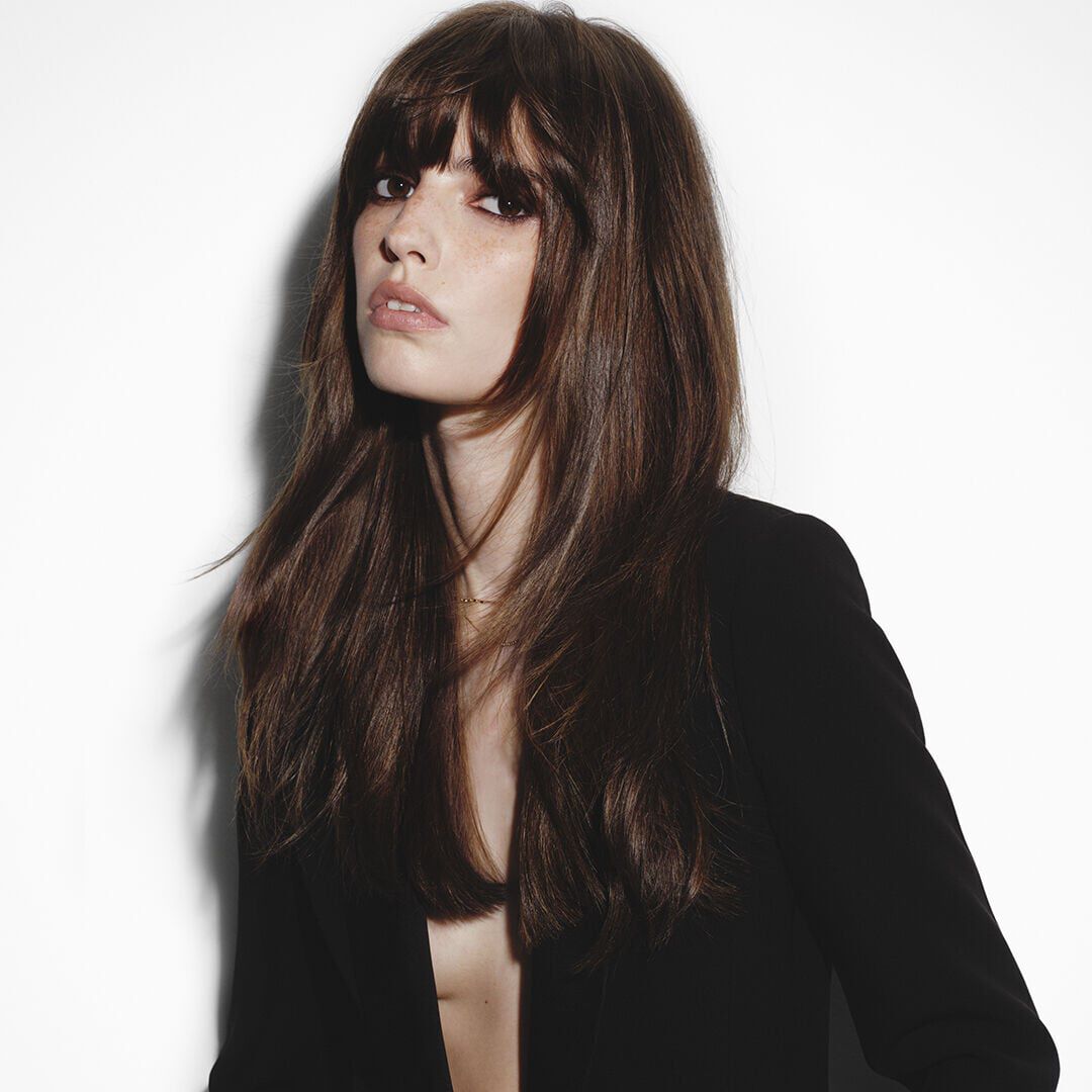 Brunette woman with bangs and long silky hair in a black blazer.