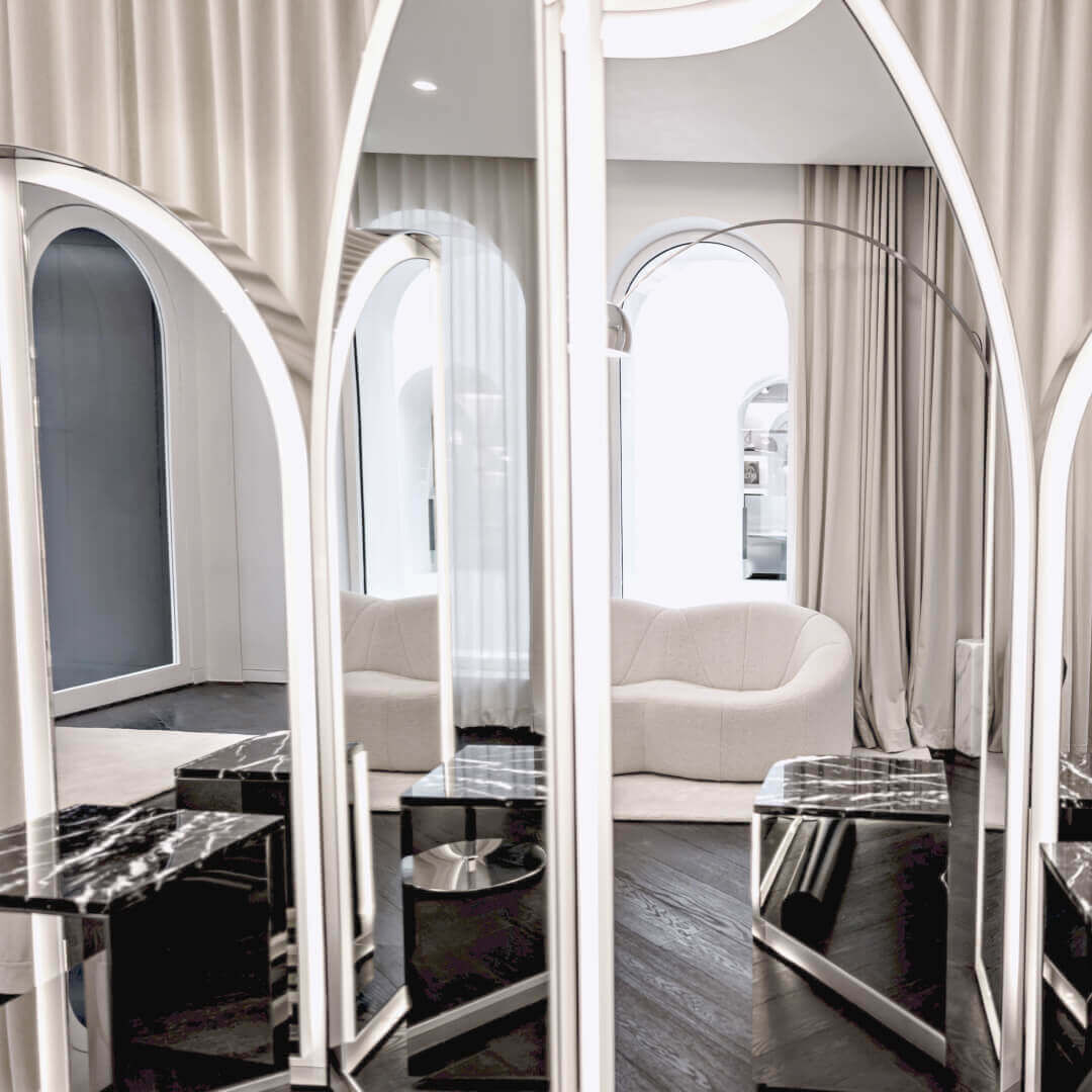 This elegant modern interior features arched reflective mirrors with linear lighting. Black marble tables and a dark wood floor are visible. A light sofa, beige curtains, and an arc lamp create a refined ambiance.