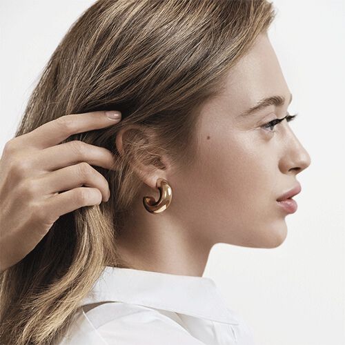 Profile portrait of a woman with light brown hair, hand adjusting a strand near her ear. She wears a white shirt and a golden hoop earring. Soft lighting highlights her hair and skin against a clean, bright background.