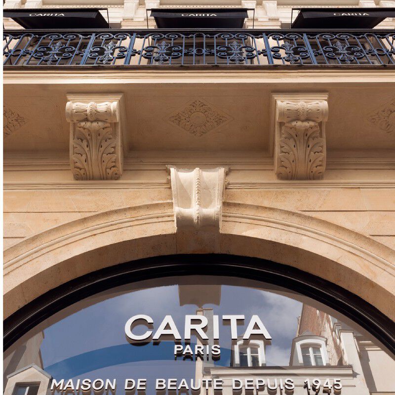 Elegant facade of the Carita Paris House. A wrought-iron balcony, sculpted architectural details, and the Carita Paris logo on the curved shop window are visible. Brand "Maison de Beaute Depuis 1945".
