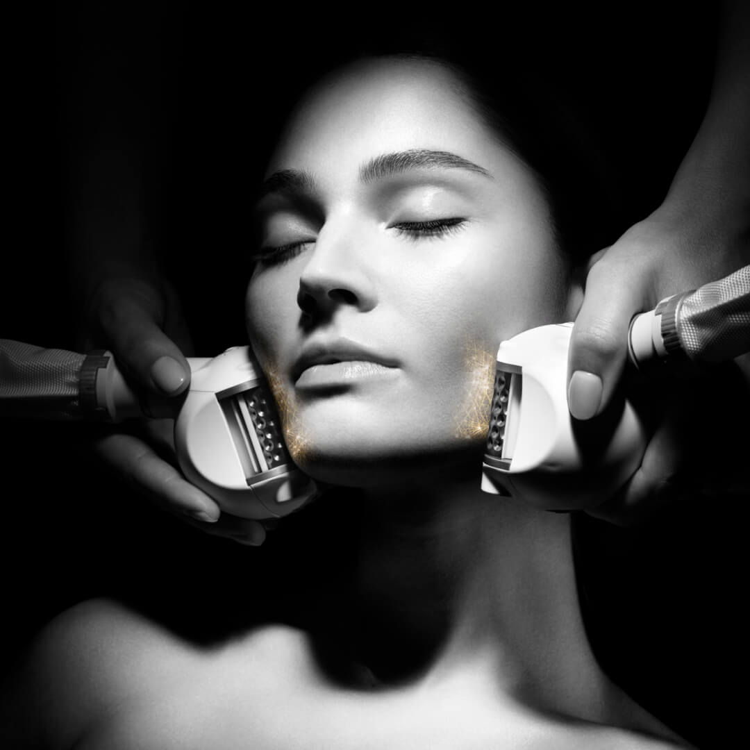 A woman receives a facial treatment with specialized devices, hands gently grasping her cheeks in a dimly lit setting.